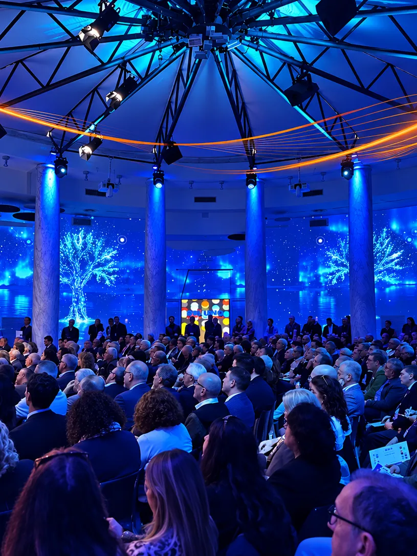 Una sala gremita sotto nastri di luce arancione: in fondo il palco con un pannello colorato e pareti blu.