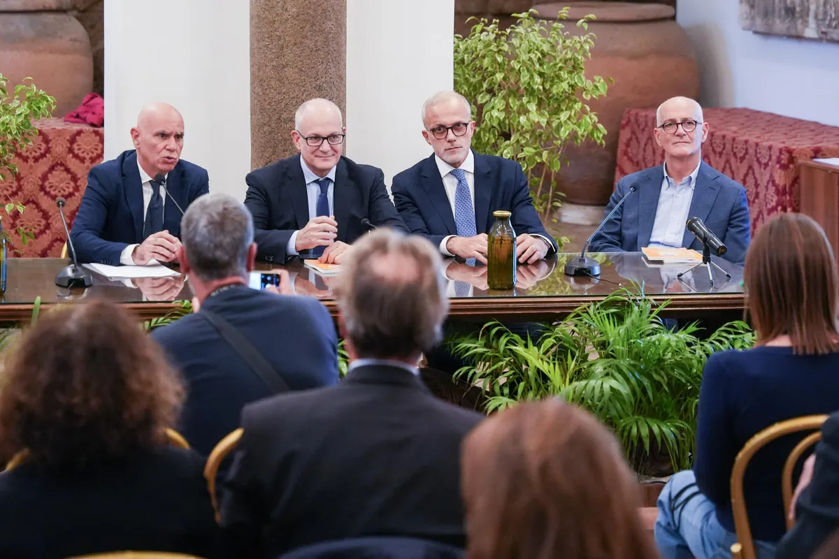 Quattro uomini al tavolo dei relatori, con i microfoni, davanti al pubblico e a un fotografo, nella sala della presentazione in Campidoglio.